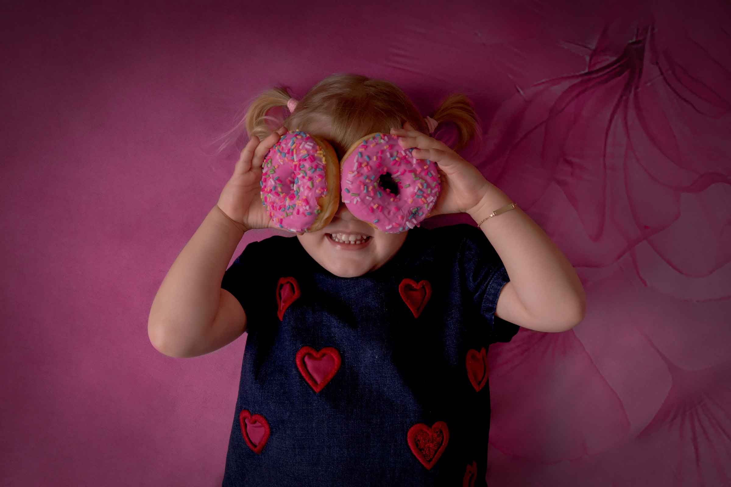 Mirkaphotography hat Blondes Mädchen im roser Hintergrund mit zwei donuts vor den Augen Fotografiert