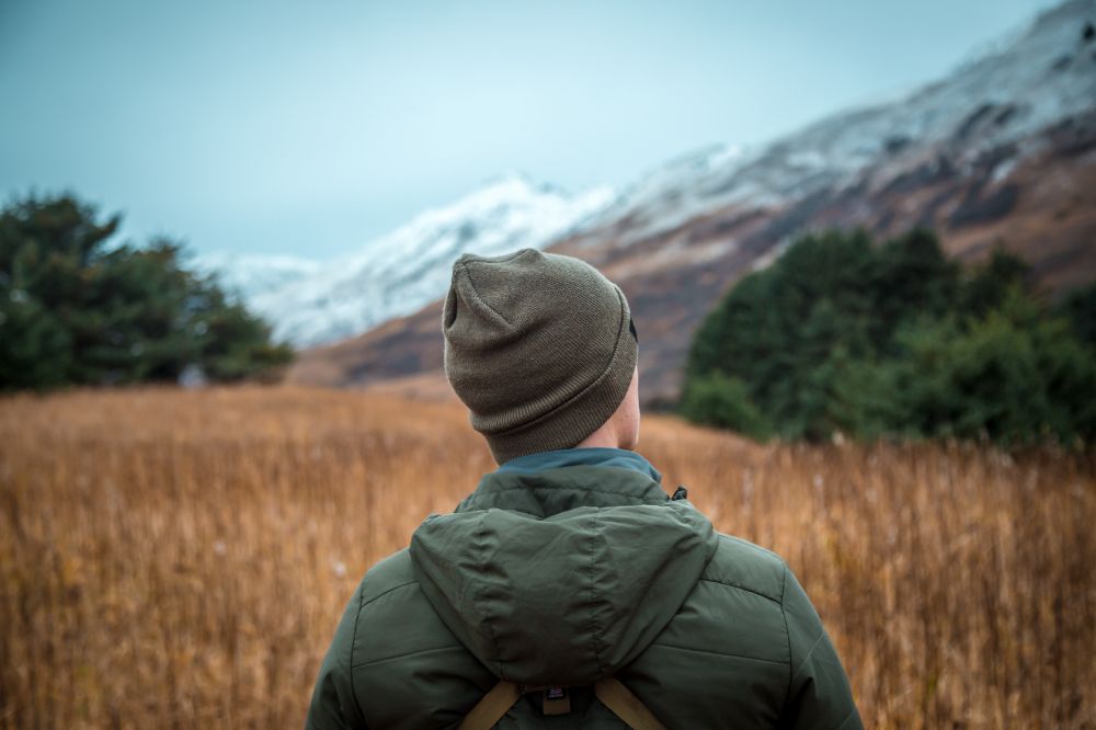 A person wearing a cap and a winter jacket, looking out at snow-covered mountains in the distance